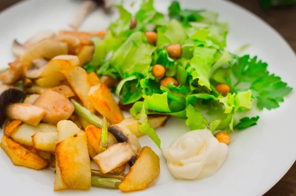 Patate fritte arrosto con funghi selvatici su piatto bianco e lattuga verde su sfondo tavolo di legno. Sfondo in legno. Vista dall'alto. Primo piano — Foto Stock