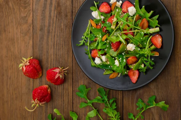 Ensalada con rúcula, fresas, requesón, aceite de oliva, sobre un plato negro, fondo de madera. Primer plano. Vista superior — Foto de Stock