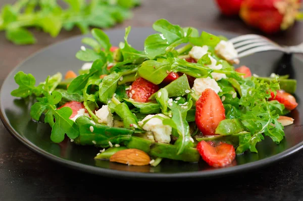 Salat mit Rucola, Erdbeeren, Quark, Olivenöl auf einem schwarzen Teller, altem schwarzem Hintergrund. Nahaufnahme. Ansicht von oben — Stockfoto