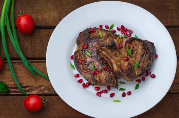 Steak pada tulang rusuk, dimasak di atas panggangan dengan buah delima di atas piring putih. Latar belakang Wooden Rustic. Tampilan atas — Stok Foto