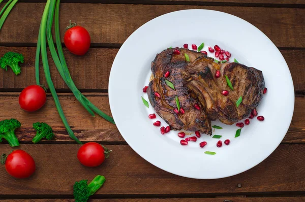 Bife em costelas, cozido em uma grelha com romã em um prato branco. Fundo rústico de madeira. Vista superior — Fotografia de Stock