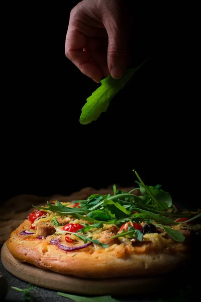 新鲜的比萨饼配西红柿, 奶酪, 鸡肉和蘑菇。意大利比萨洒了芝麻菜, 用手累了。黑色乡村背景。特写 — 图库照片