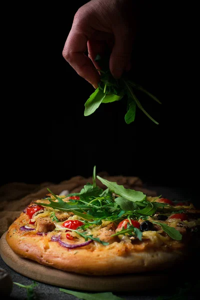 Pizza proaspătă cu roșii, brânză, pui și ciuperci. Pizza italiană presărată cu arugula, obosită de muncă cu mâinile. Fundal rustic negru. Closeup — Fotografie, imagine de stoc