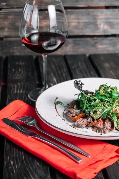 Koyu ahşap arka plan üzerinde kırmızı peçete ile kadeh şarap ve sıcak Dana ile salata servis