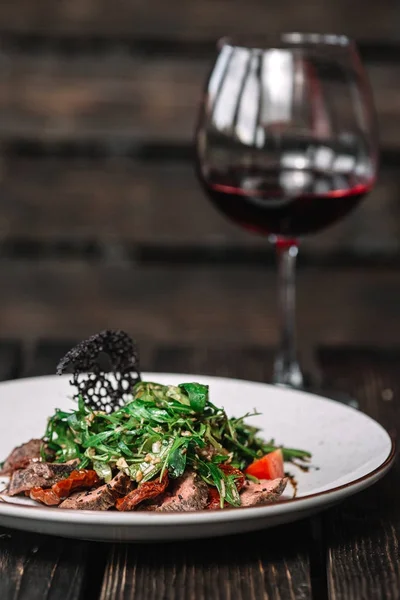 Стакан вина и салат с теплой телятиной, сушеными помидорами и рукколой на темном деревянном фоне — стоковое фото