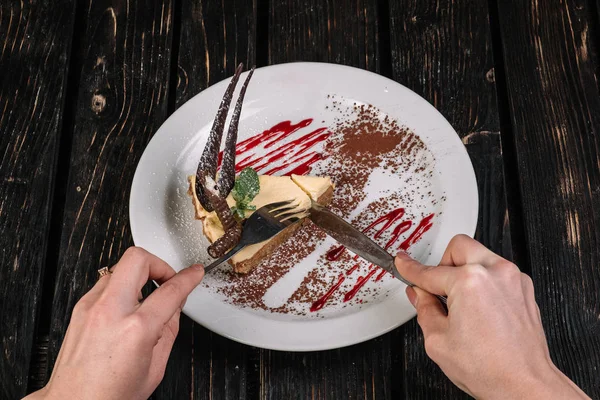 Hands holding knife and fork in front of plate with cheese cake on dark wood background.