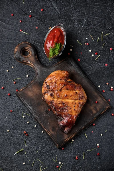 Grilled chicken fillets on wooden cutting board with tomato sauce. Black concrete background. Top view
