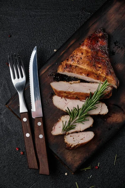 Grilled chicken fillets on wooden cutting board with rosemary and cutlery on black background. Top view