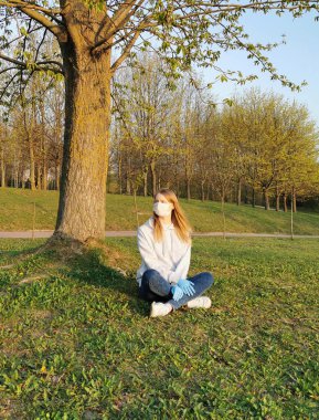 İlkbaharda bir ağacın altında maske ve eldiven takan bir kız. Coronavirus Koruma 2020.
