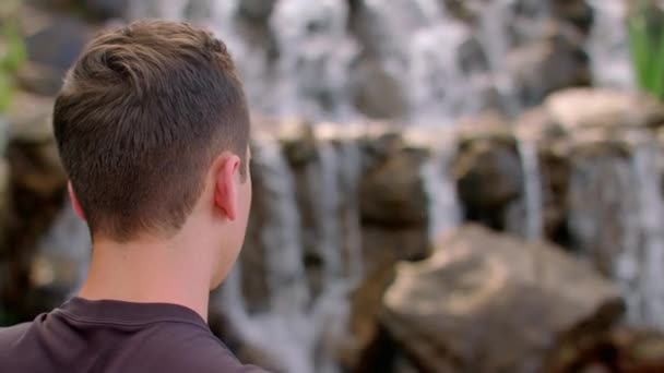 Young man looking at mountain waterfall. Hiker resting near mountain waterfall — Stock video