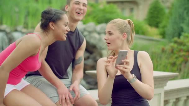 Веселые друзья веселятся в парке. Молодые люди используют телефон на открытом воздухе летом — стоковое видео
