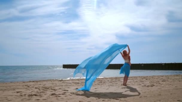 Женщина с беременным животом, держащая голубую ткань, летящую на ветру на пляже — стоковое видео