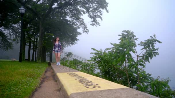 Fille souriante marchant sur parapet dans le parc. Femme s'approchant de la mer — Video