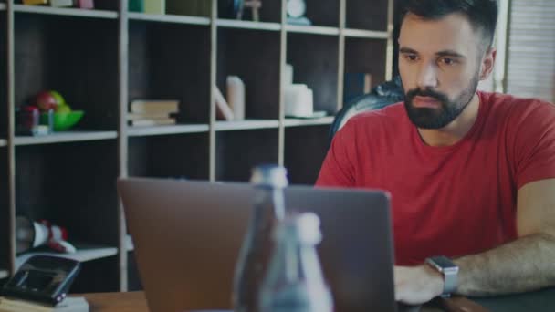 Бородач работает на ноутбуке в домашнем офисе. Человек смотрит экран ноутбука — стоковое видео