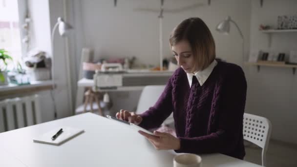 Mujer joven usando tableta de computadora sentada en la mesa en casa — Vídeos de Stock