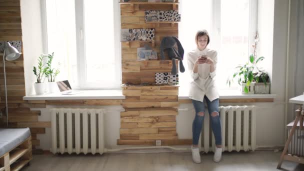 Young woman holding mobile phone in hands and standing front window — Stock Video