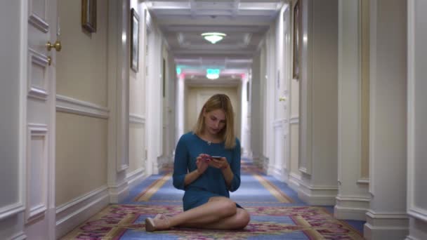 Молода жінка сидить на середньому поверсі в довгому коридорі і використовує мобільний телефон — стокове відео