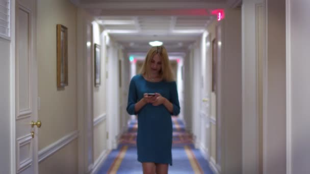 Νεαρή γυναίκα το περπάτημα σε μακρύ διάδρομο στον διάδρομο και ψάχνει για smartphone οθόνη — Αρχείο Βίντεο