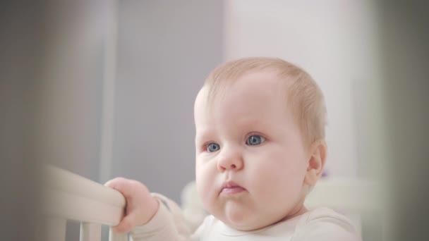Linda cara de bebé con ojos azules. Bebé de pie en la cama y mirando a su alrededor — Vídeo de stock
