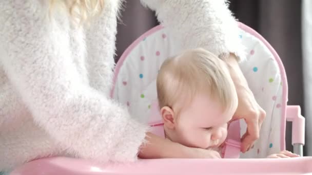 Madre poniendo al bebé en la silla en la cocina. Mamá manos abrochan cinturones en silla rosa — Vídeos de Stock