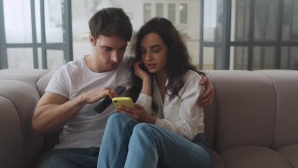 Pareja feliz sentada en un sofá con teléfonos móviles juntos. Hombre y mujer sonrientes — Vídeos de Stock