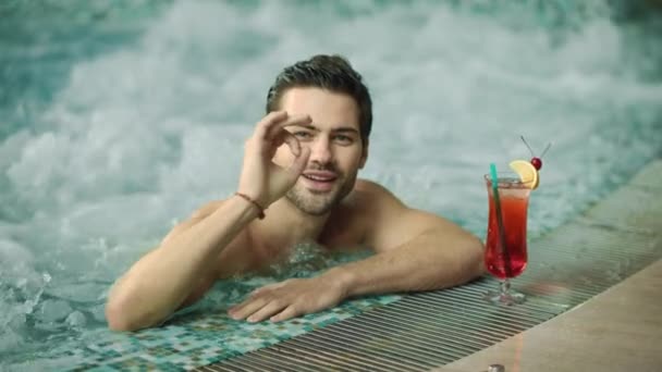Macro of smiling guy making ok gesture. Man posing for camera at luxury pool. — Stock Video