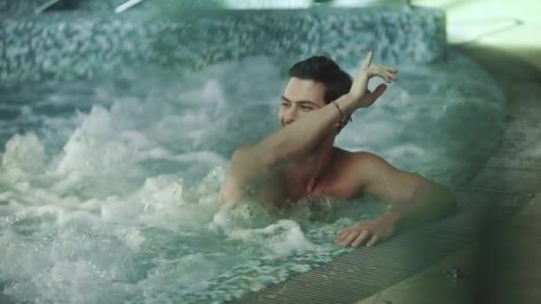 Homem alegre descansando no banho de hidromassagem. Homem sorridente dizendo oi no hotel spa. — Vídeo de Stock