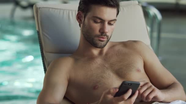 Un homme qui regarde le téléphone près de la piscine. Homme relaxant avec téléphone au centre de bien-être — Video