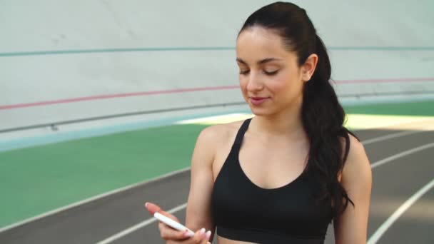 Πορτρέτο του αθλητισμού γυναίκα χρησιμοποιώντας το τηλέφωνο στο γήπεδο. Γυμναστική γυναίκα ψάχνει smartphone — Αρχείο Βίντεο