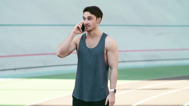 Sportovní muž volat mobilní telefon stojící na trati venkovní. Fitness muž mluví telefon — Stock video