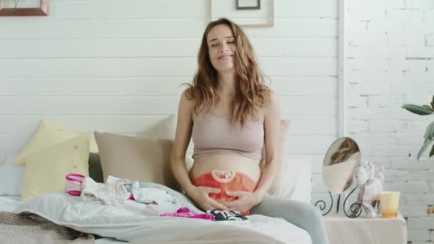 Barriga sorridente mãe segurando roupas de bebê na cama. Mulher grávida sentado quarto — Vídeo de Stock