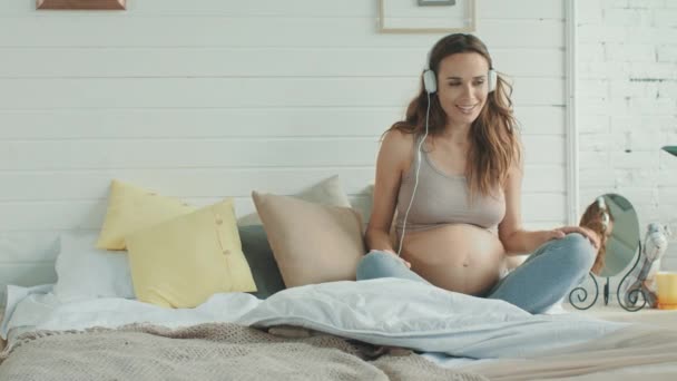 Lächelnde werdende Mutter mit Kopfhörern auf nacktem Bauch im Schlafzimmer. — Stockvideo