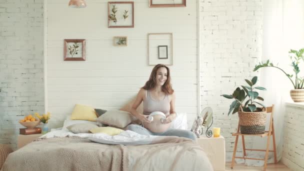 Mujer embarazada que aplica auriculares en el vientre. Embarazada escuchando música en la cama . — Vídeos de Stock