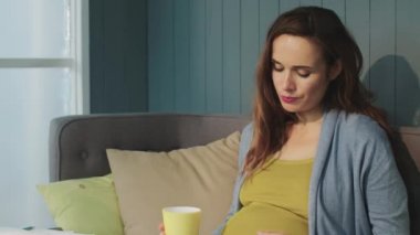 Macro pregnant woman drinking from glass mug in living room.
