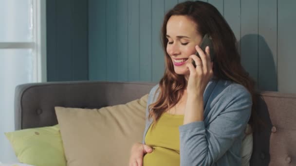 Портрет усміхненої вагітної жінки, що говорить смартфон на дивані в сучасному будинку . — стокове відео