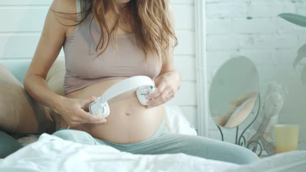 Unrecognized pregnant woman holding headphones on her belly at home. — Stok video