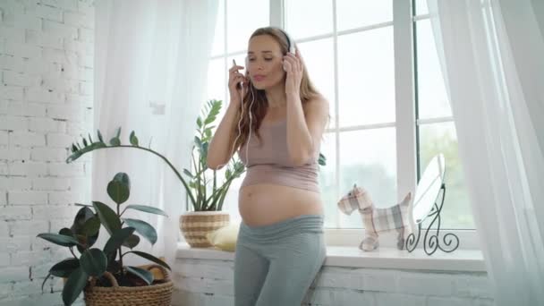 Lächelnde werdende Mutter, die über Kopfhörer Musik hört. Schwangere tanzen zu Hause — Stockvideo