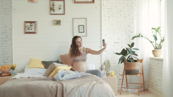 Gülümseyen hamile kadın selfie çekiyor. Müstakbel anne yatakta oturuyor.. — Stok video