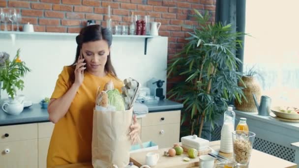 Femme ramenant des sacs d'épicerie à la maison. Femme au foyer finissant appel téléphonique dans la cuisine . — Video