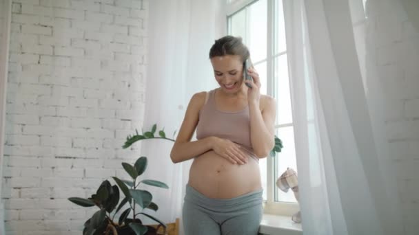 Беременная женщина разговаривает по телефону дома. Ожидаемая мать гладит голый живот — стоковое видео