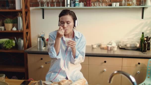 Жінка їсть йогурт на кухні. Красива дівчина слухає музику в навушниках . — стокове відео