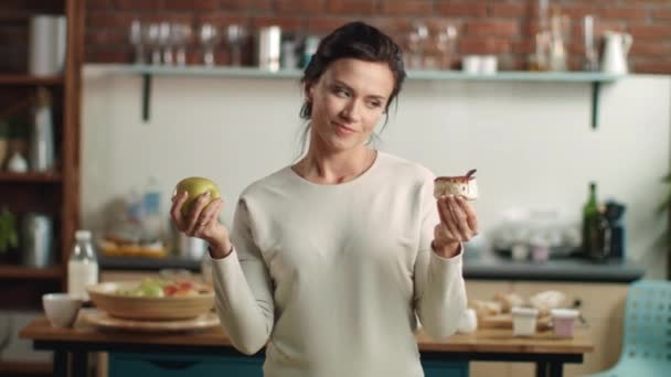 Mulher fazendo escolha entre maçã e bolo na cozinha. Menina escolhendo dieta de frutas . — Vídeo de Stock