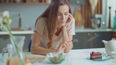 Thoughtful woman choosing between cake and salad on kitchen