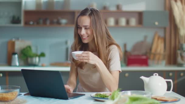 Женщина ведет видео чат на ноутбуке на кухне. Девушка пьет кофе дома — стоковое видео
