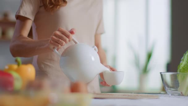 Egy nő teát tölt csészébe a konyhában. Egy hölgy teát iszik otthon. — Stock videók