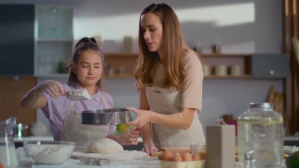 Woman teaching daughter to sieve flour on dough at kitchen — Stock video