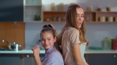 Happy daughter and mother dancing back to back on kitchen in slow motion