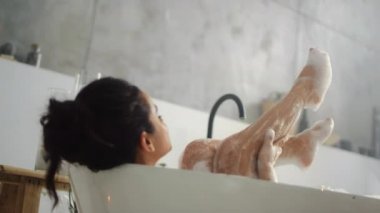 Köpük banyosunda bacaklarını yıkayan romantik bir kadın. Küvette bacakları temizleyen esmer kız.