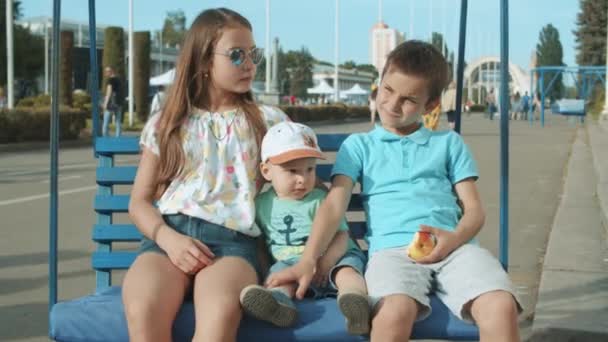 Entspannte kleine Kinder sitzen auf einer Schaukel. Nette drei Geschwister verbringen Zeit im Freien — Stockvideo