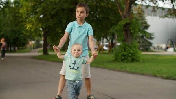 Az idősebb testvér segít Juniornak kimenni a szabadba. Családi séta a nyári parkban — Stock videók